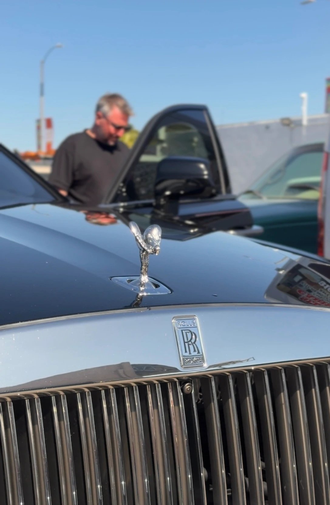 A man at the open door of a dark Rolls-Royce, seen over the car's chrome grille and hood ornament in bright daylight